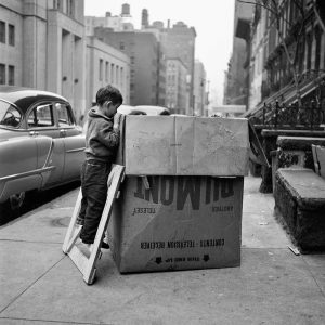 تصویری از عکس خاکستری ویویان مایر که در آن پسر بچه‌ای در پیاده رو روی تکه چوبی ایستاده و داخل جعبه را تماشا می‌کند